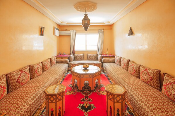 A traditional Moroccan room with a central seating area, a chandelier, and a large window with curtains.