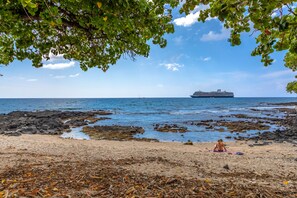 A beach with a ship in the distance, rocky shoreline, and a person sitting on a towel. - Kona Islander Inn - Prime Downtown Location with Hot Tub, Pool & Beach! (Kailua-Kona)