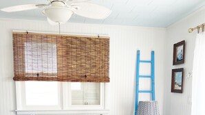 A room with a blue sofa, a white ceiling fan, a window with bamboo blinds, a blue ladder, and a small table with a lamp.