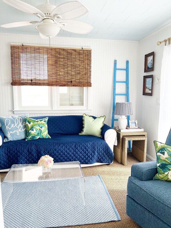 A room with a blue sofa, a white ceiling fan, a window with bamboo blinds, a blue ladder, and a small table with a lamp.