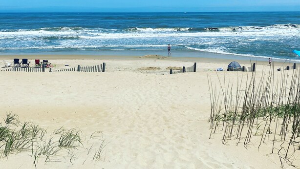 A sandy beach with a clear blue sky, waves in the ocean, and a few beach chairs and umbrellas.