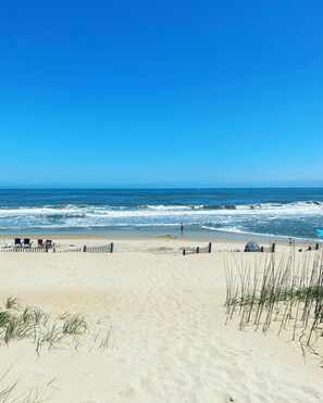 A sandy beach with a clear blue sky, waves in the ocean, and a few beach chairs and umbrellas.