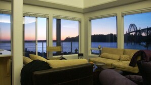 A living room with a large window overlooking a body of water and a bridge. There is a sofa and a small table with a lamp.