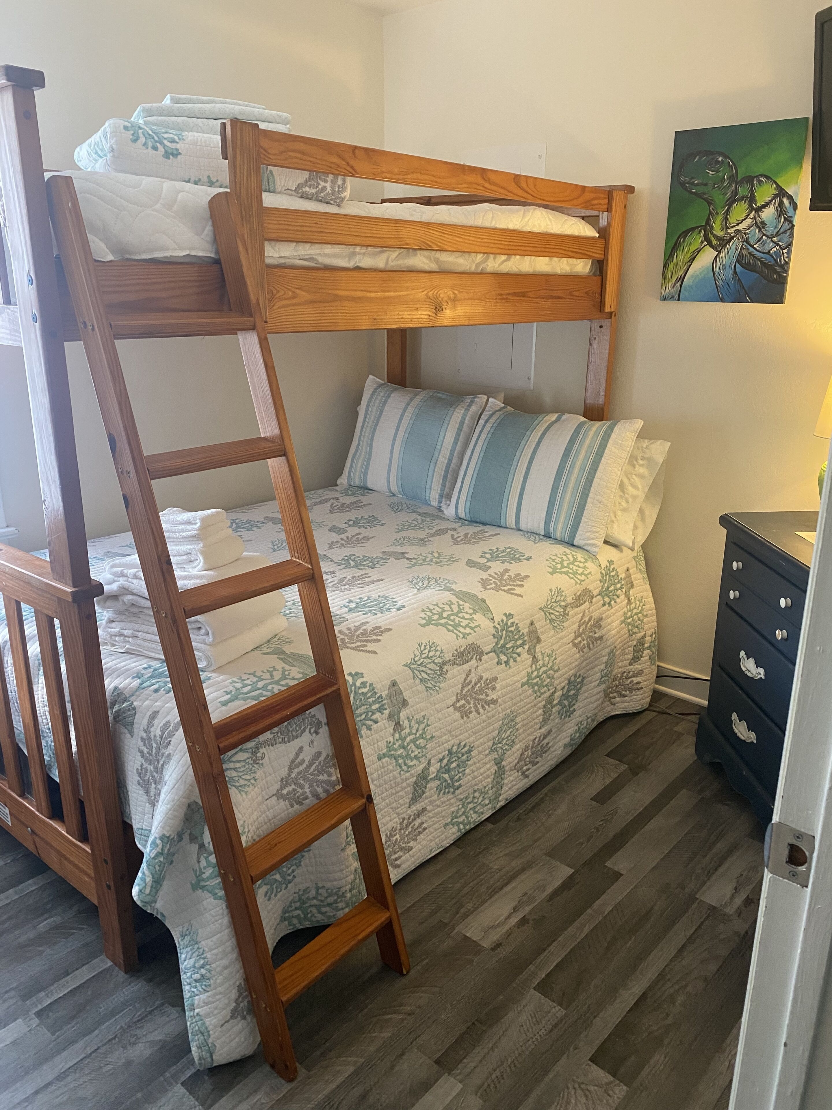 A bunk bed with a wooden ladder, a patterned bedspread, and pillows. There is a dresser with drawers and a painting on the wall.