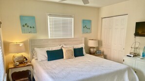 Une chambre à coucher bien rangée, avec un grand lit, deux tables de chevet, une commode, un téléviseur fixé au mur et une porte.