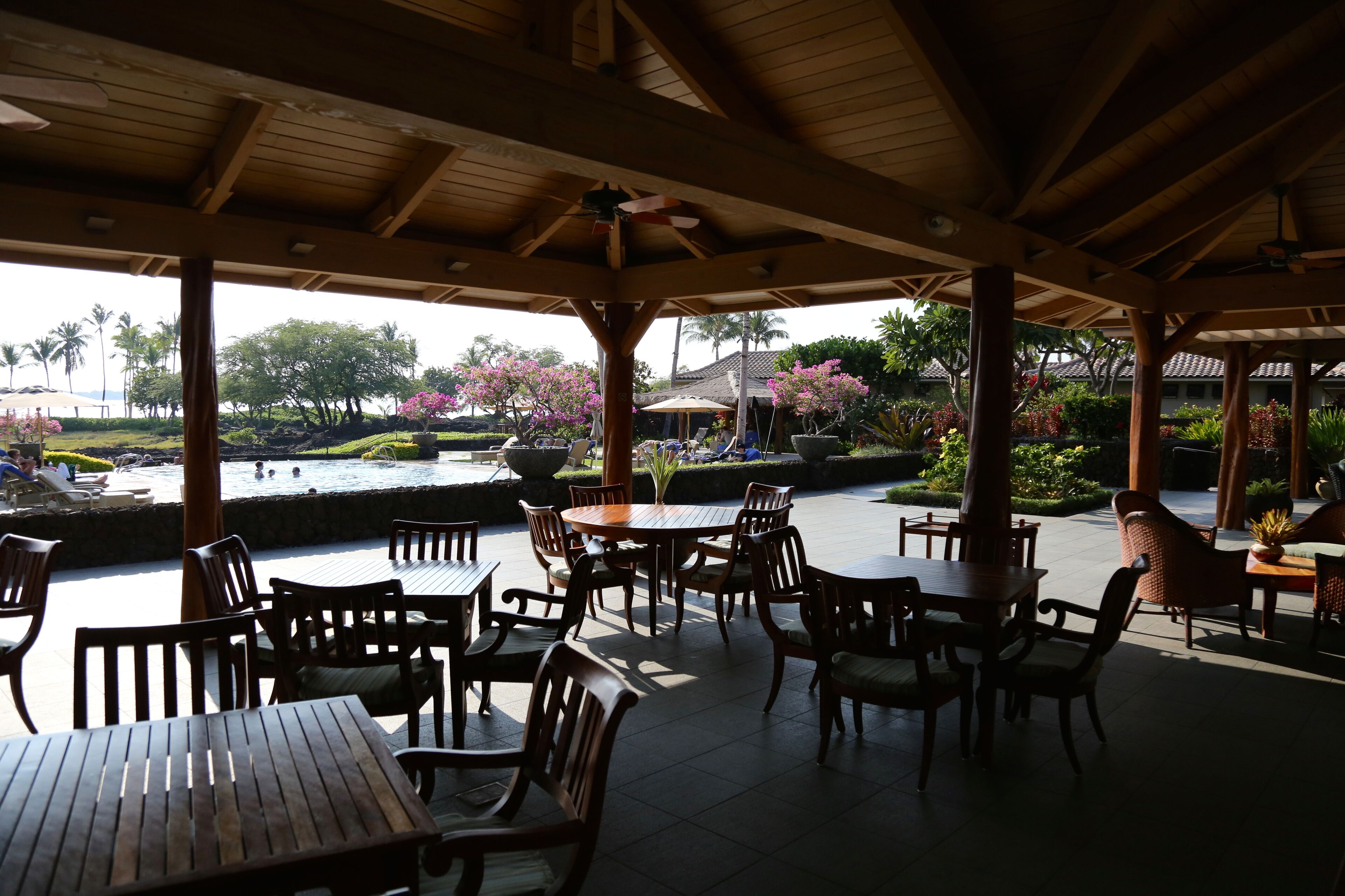 Un patio exterior con mesas y sillas de madera, rodeado de exuberante vegetación y vistas a una piscina.