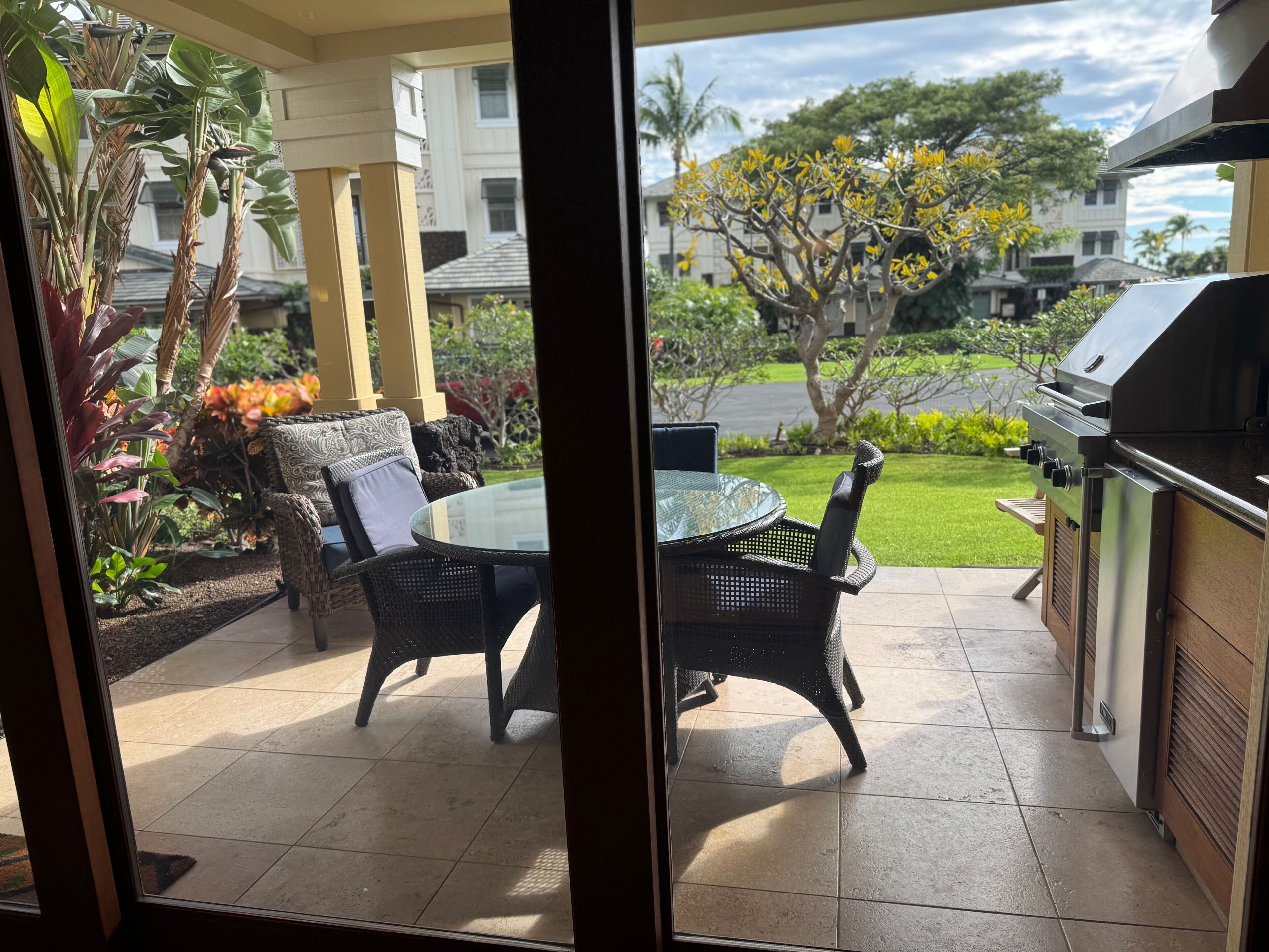 A patio with a glass table and wicker chairs, a grill, and a view of a garden and buildings.