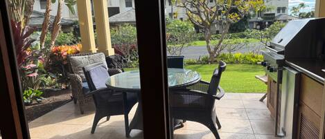 A patio with a glass table and wicker chairs, a grill, and a view of a garden and buildings.