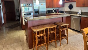 Una cocina con armarios de madera, electrodomésticos de acero inoxidable, encimera de granito y dos taburetes de mimbre.