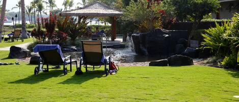 A tropical resort setting with a thatched-roof gazebo, a small pond, and lounge chairs on a grassy area.