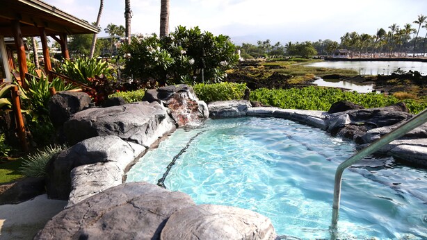 Un jacuzzi rodeado de rocas y plantas tropicales, con vista a un lago y palmeras al fondo.