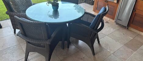 Une table ronde en verre, des chaises en osier, un barbecue et une plante en pot.