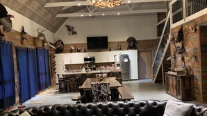 Un vaste espace de vie comprenant un grand canapé d’angle en cuir, une table basse en bois et un lustre moderne. On aperçoit à l’arrière-plan une cuisine avec des meubles blancs et un coin repas avec une table et des chaises.