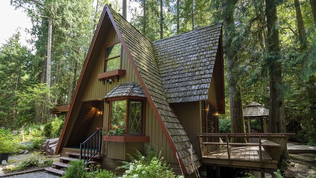 A cabin with a steeply pitched roof, a wooden deck, and a staircase leading to a second-story window. - Charming Chalet-Private Cedar Soaking Tub-Snowline-Owner Managed (Glacier)