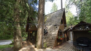 A wooden cabin with a sloped roof, surrounded by tall trees. There is a deck with outdoor seating and string lights. - Charming Chalet-Private Cedar Soaking Tub-Snowline-Owner Managed (Glacier)