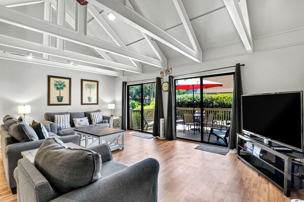 A spacious living area with a sofa set, a coffee table, a flat-screen TV, and a sliding glass door leading to an outdoor patio.