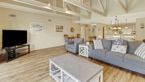 A spacious living room with a large flat-screen TV, a grey sofa set, and a coffee table. There are framed pictures on the walls and a kitchen area visible in the background.