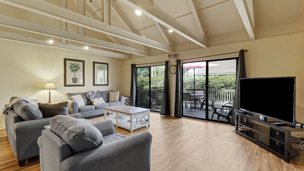 A spacious living room with a grey sofa, a coffee table, and a TV. There is a balcony with a table and chairs.