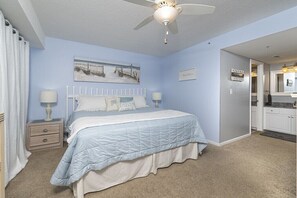 Une chambre à coucher avec un lit, des tables de chevet, un ventilateur de plafond, un tableau et une vue sur la cuisine.