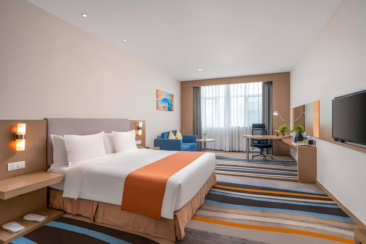 Une chambre d’hôtel avec un grand lit, un bureau avec une chaise, une petite table, un téléviseur et une fenêtre avec des rideaux.