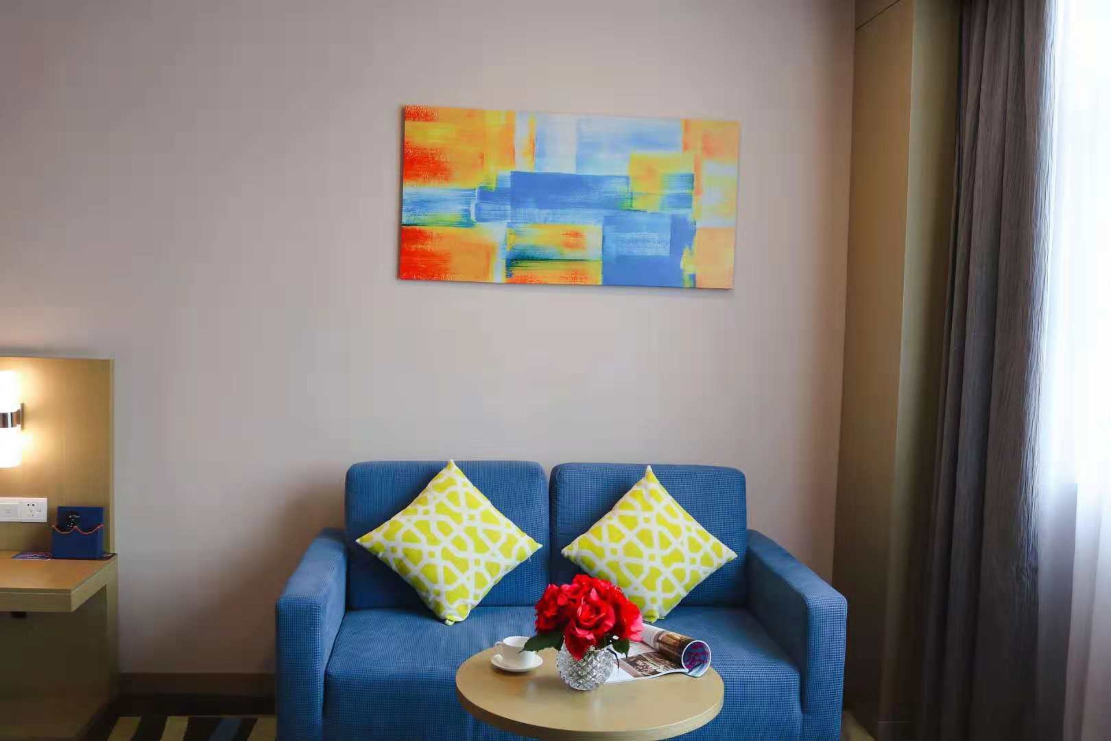 A room with a blue sofa, two patterned pillows, a small round table with a vase of red flowers, and a colorful abstract painting on the wall.