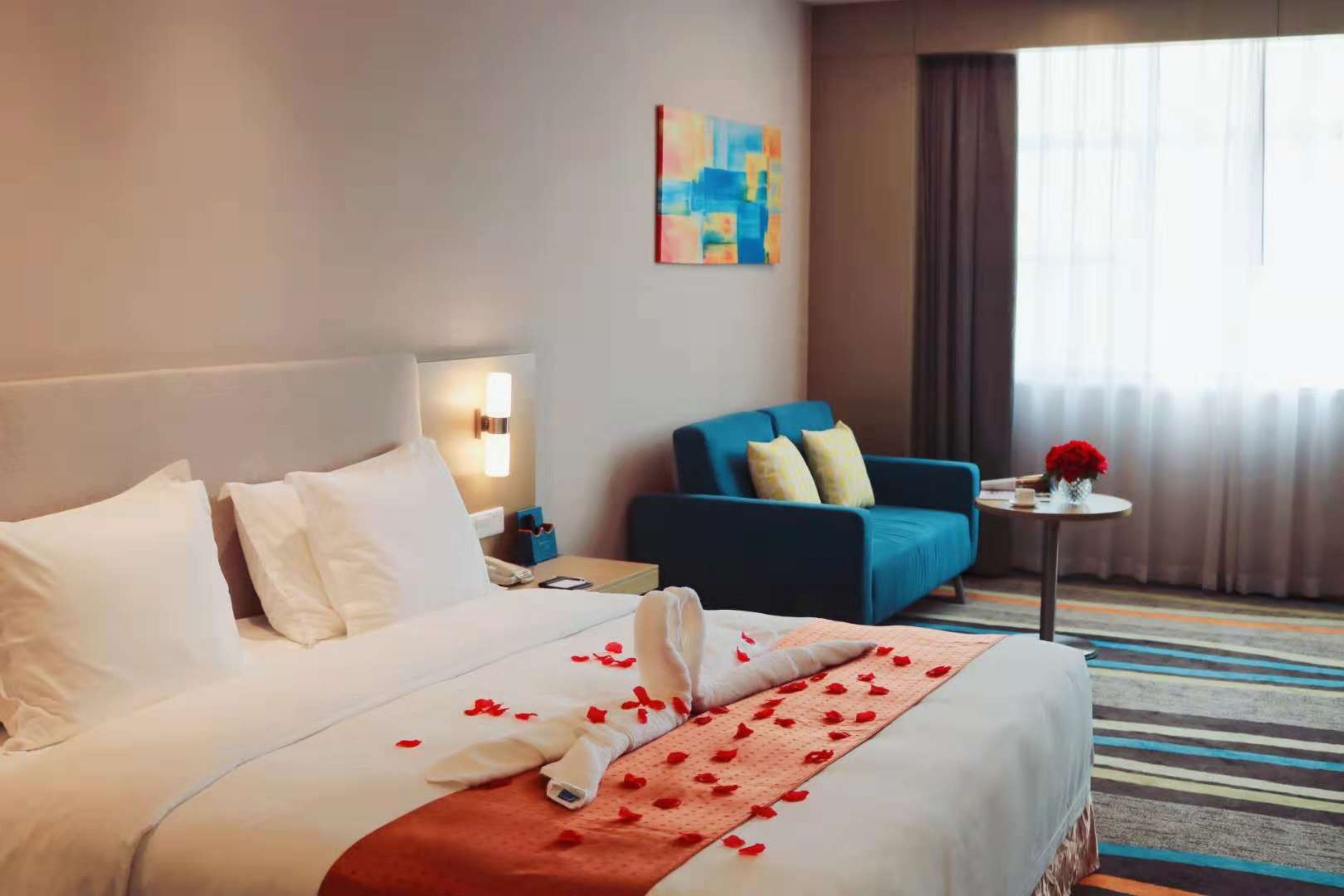 A hotel room with a large bed, a blue sofa, a small table with a vase of flowers, and a painting on the wall.