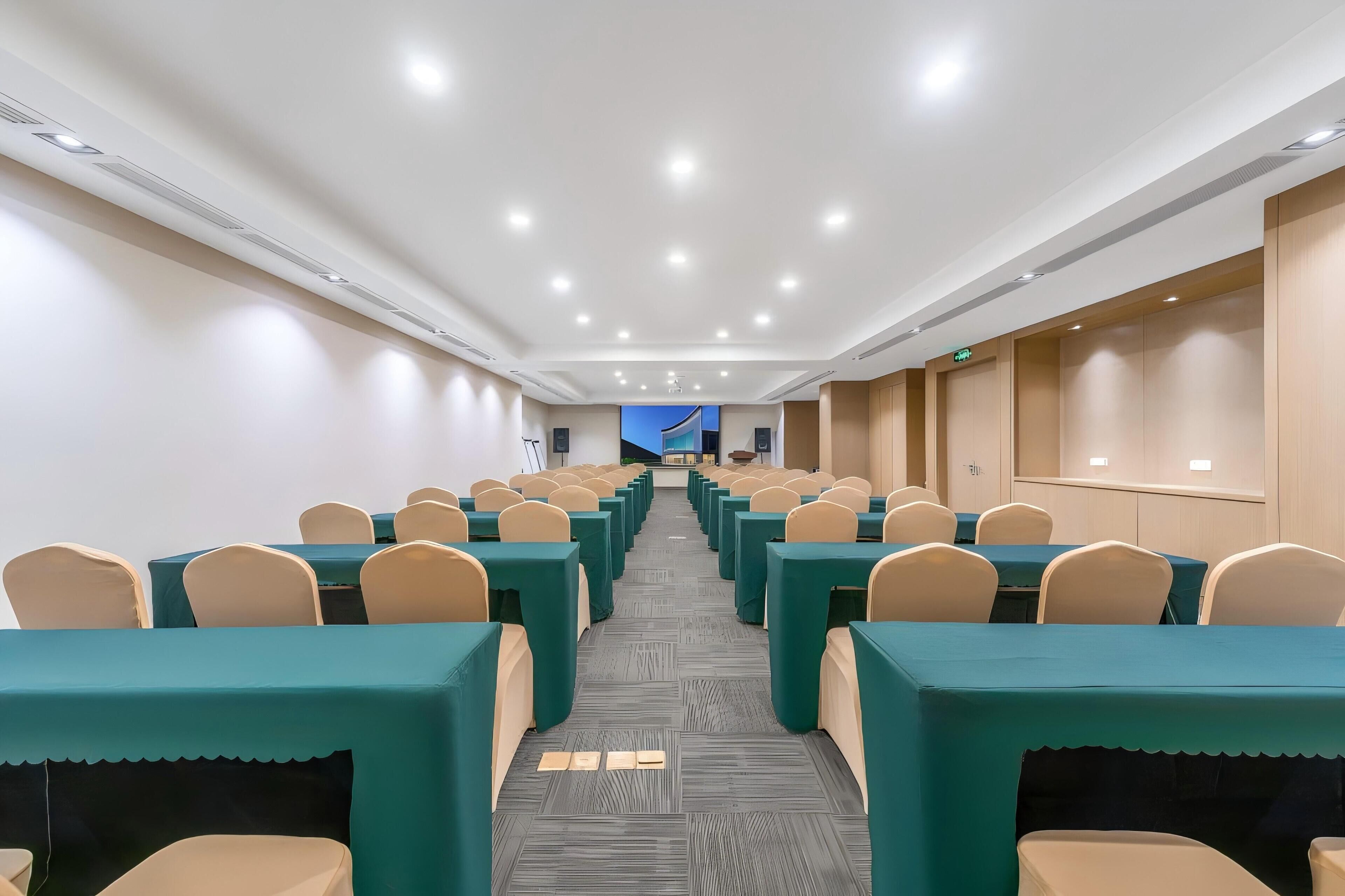 Une salle de conférence avec des rangées de tables et de chaises, un sol recouvert de moquette et un plafond moderne doté d’un éclairage encastré.