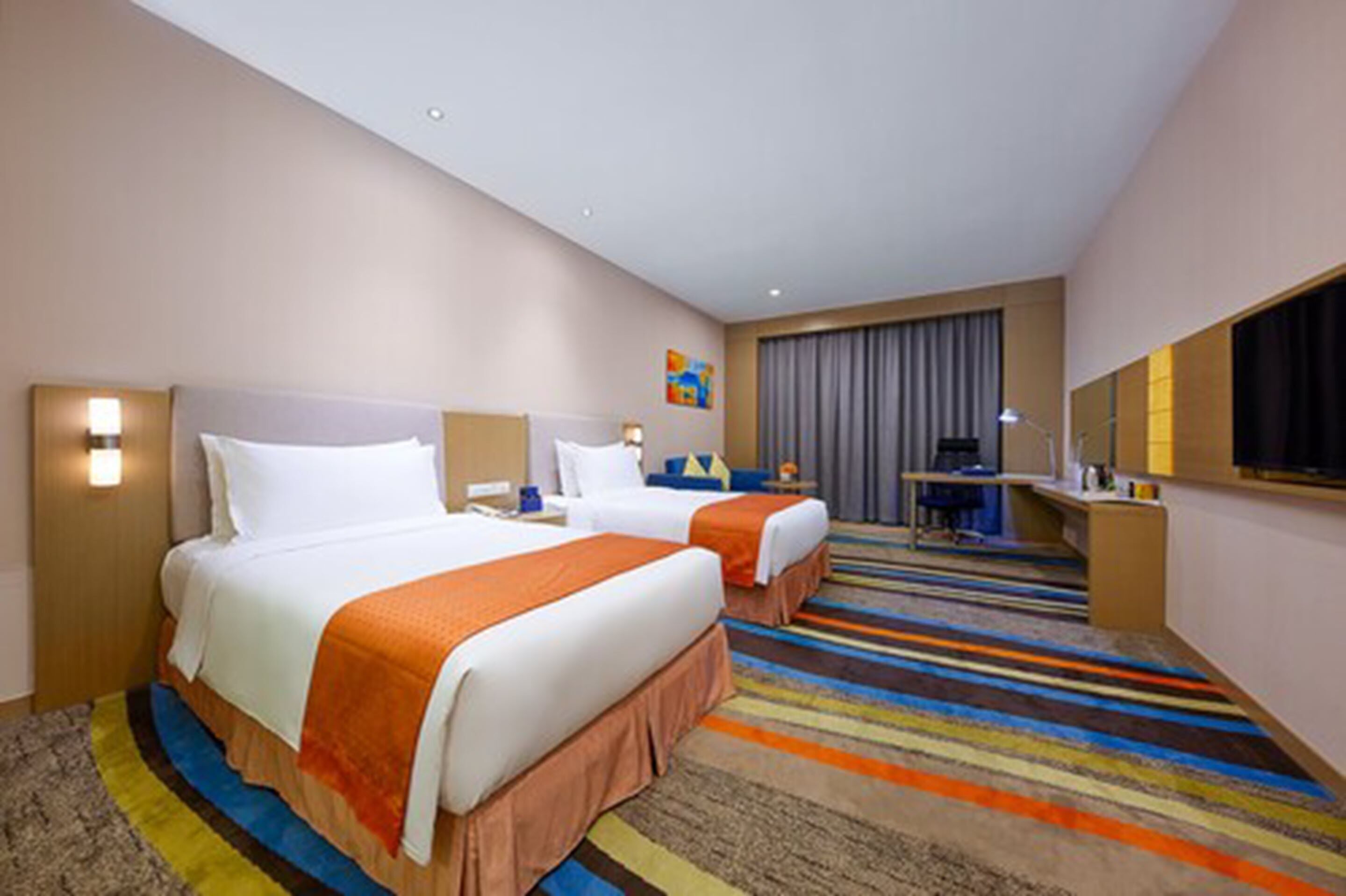 A hotel room with two beds, a desk, a chair, a TV, and a colorful striped carpet.
