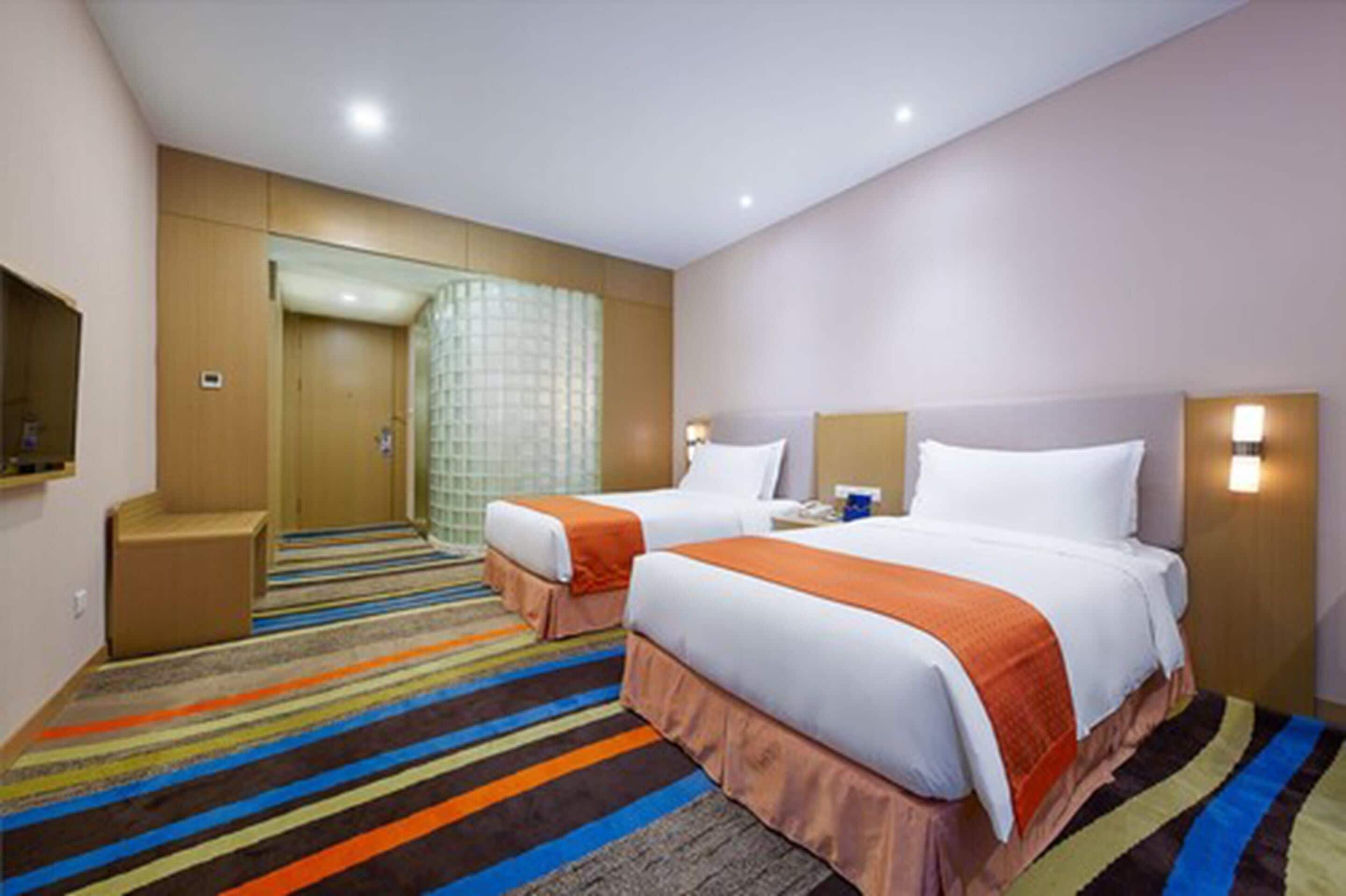 Una habitación de hotel con dos camas, una alfombra colorida a rayas, un espejo grande y un baño con cabina de ducha de vidrio.