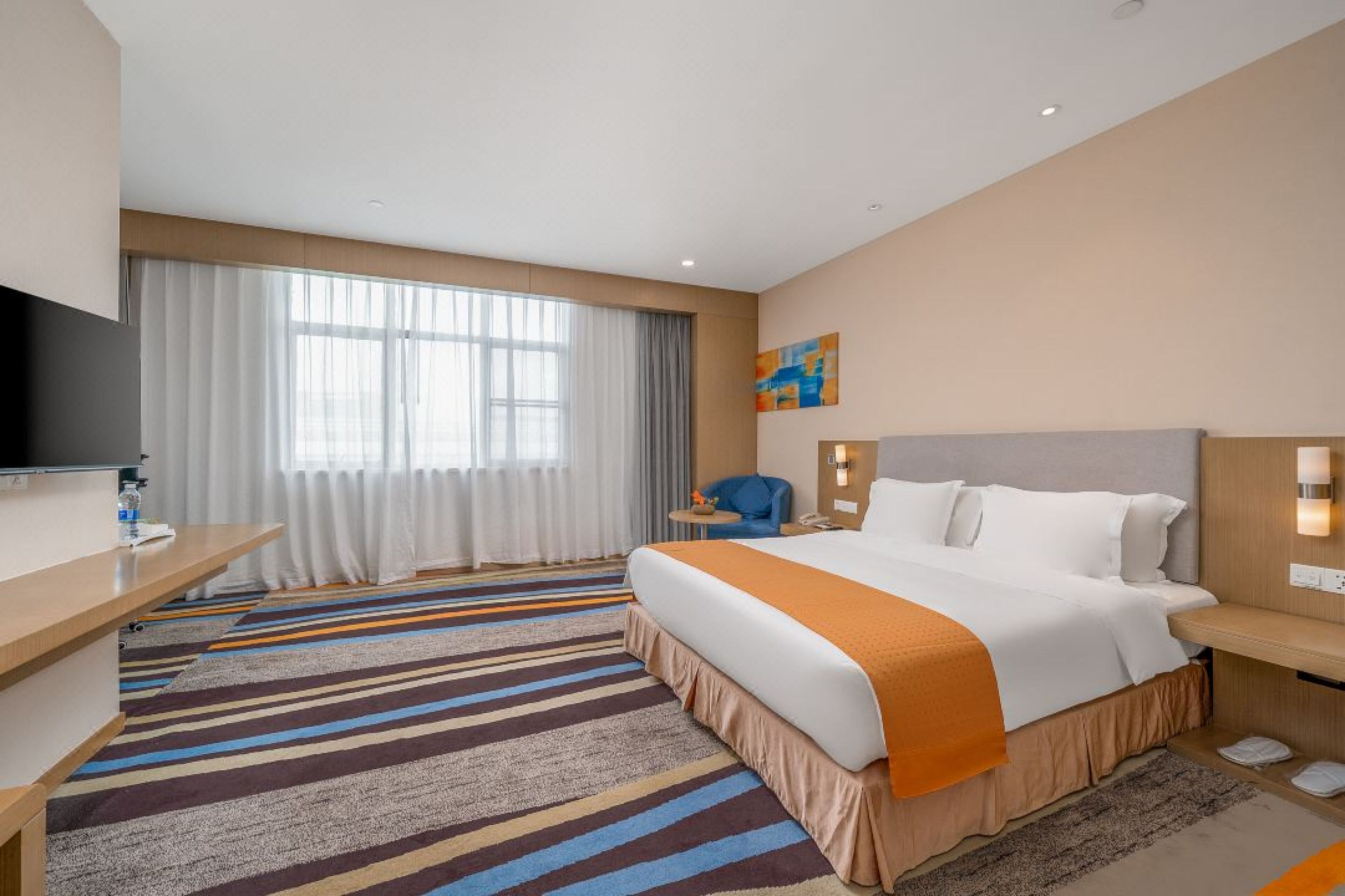 A hotel room with a large bed, a desk, a TV, and a window with curtains.