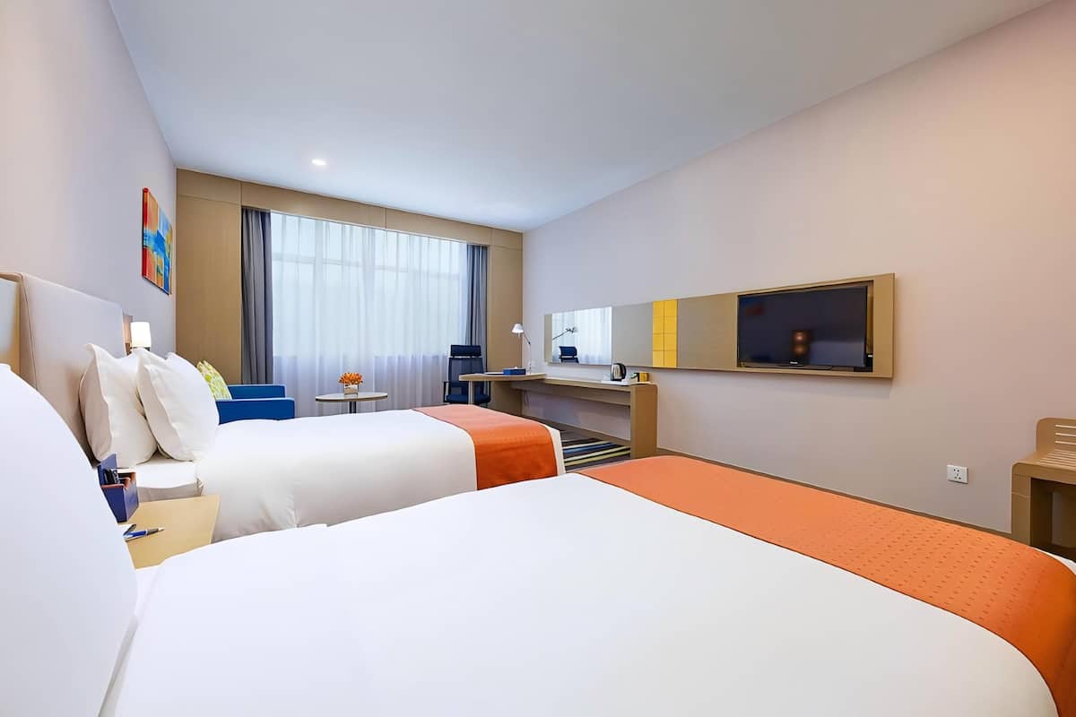 Une chambre d’hôtel avec deux lits, un bureau, une chaise, une petite table avec des fleurs, une télévision et une fenêtre avec des rideaux.