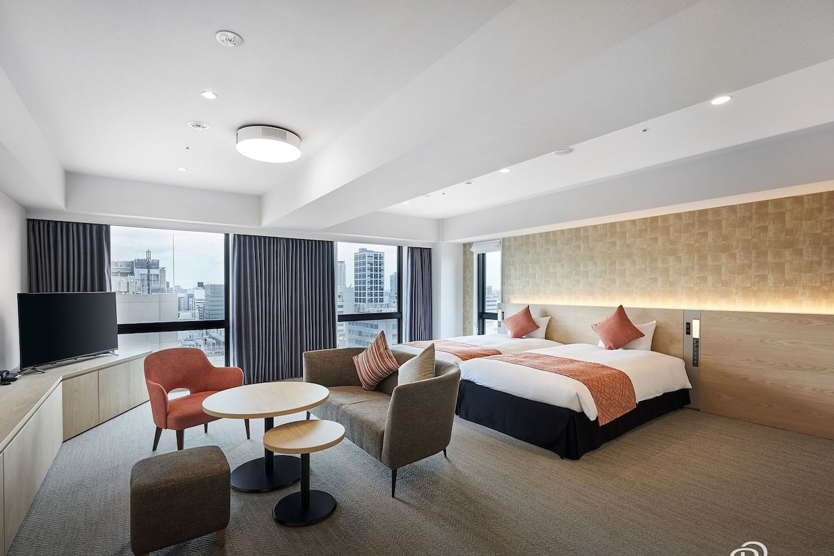Habitación de hotel con una cama grande, dos sillones, una mesita, un televisor y vistas a la ciudad.