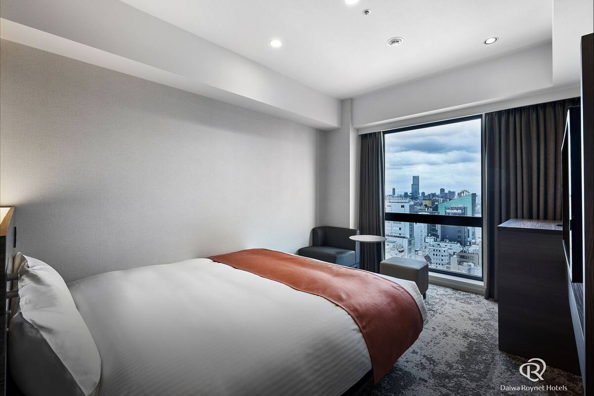 Habitación de hotel con una cama grande, un escritorio, una silla y vistas a un paisaje urbano a través de un ventanal.