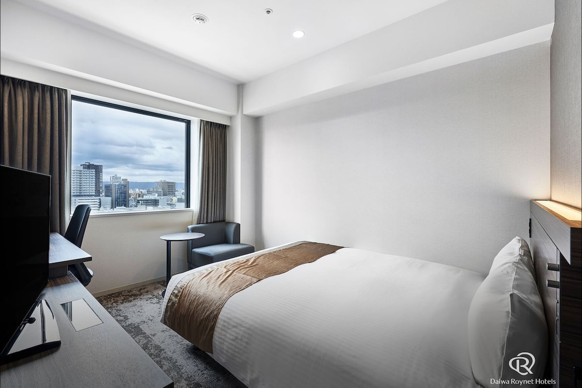 Habitación de hotel con cama, escritorio con silla, televisión y ventana con cortinas.
