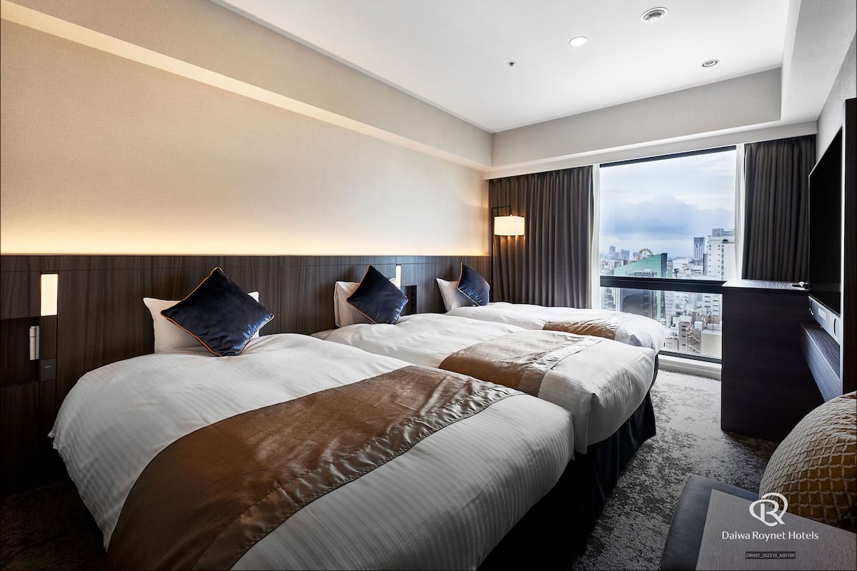 Habitación de hotel con dos camas, un ventanal grande con vistas a la ciudad, un televisor de pantalla plana y un minibar.