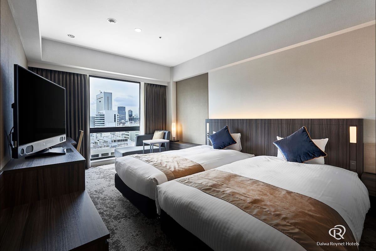 Habitación de hotel con dos camas, televisión, escritorio y vista a la ciudad.