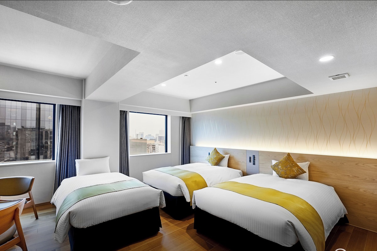 Habitación de hotel con dos camas, una silla, ventana con vista a la ciudad e iluminación empotrada en el techo.