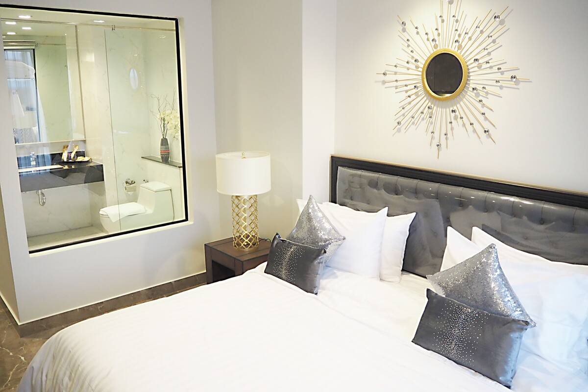 A modern hotel room with a large bed, a nightstand with a lamp, a bathroom mirror, and a decorative wall piece.