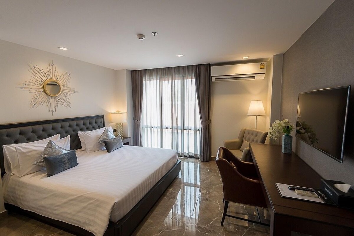 A hotel room with a large bed, a desk with a TV, a chair, and a sunburst wall decoration.