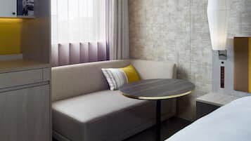 Quarto de hotel moderno com sofá, uma mesinha redonda, cama e luminária fixada na parede.