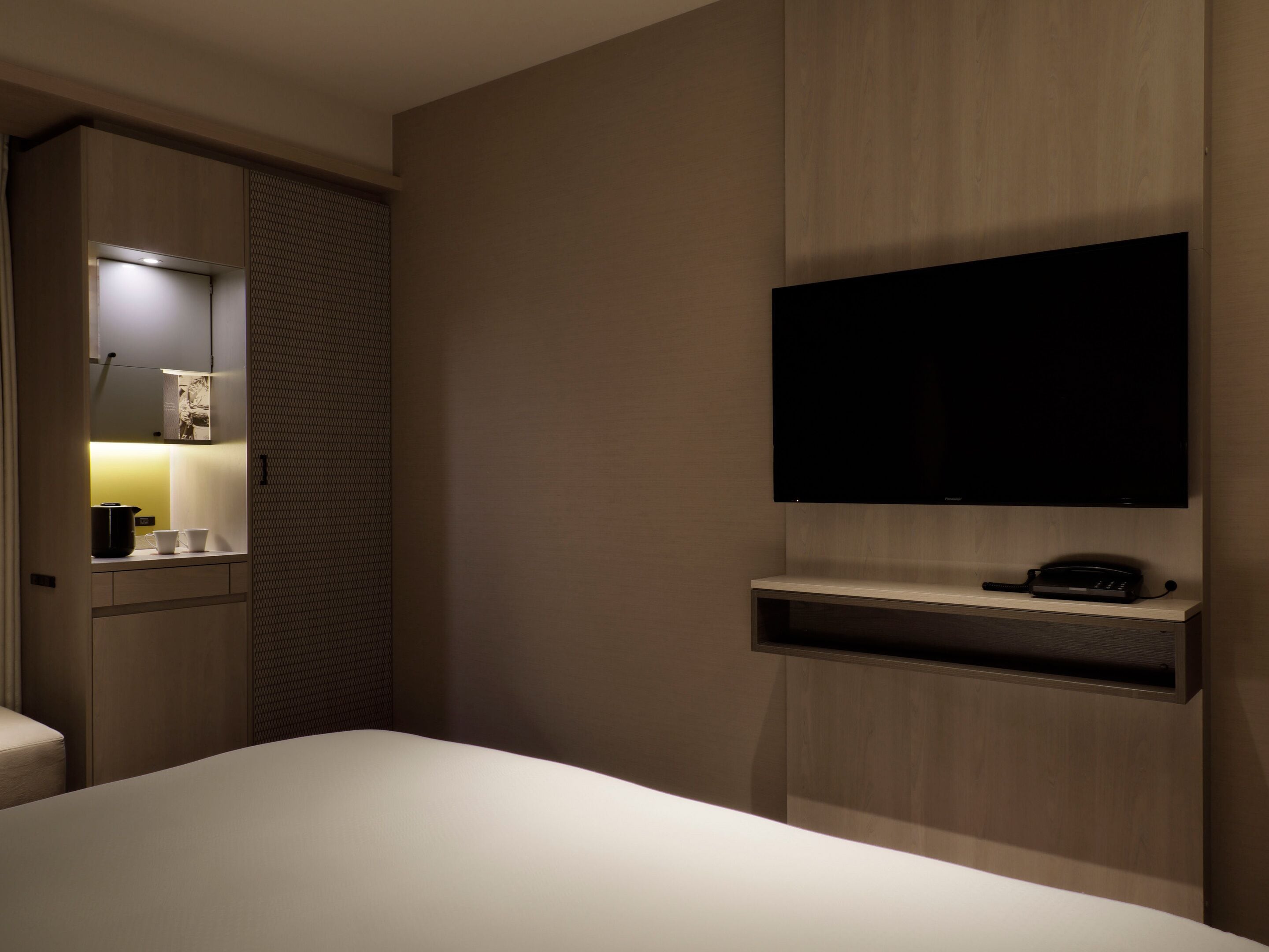 Ein Hotelzimmer mit Bett, einem an der Wand montierten Fernseher, einem kleinen Regal darunter und einem Kleiderschrank mit Spiegel und Beleuchtung.