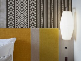 Quarto de hotel com parede estampada, sofá com almofada e luminária pendente.