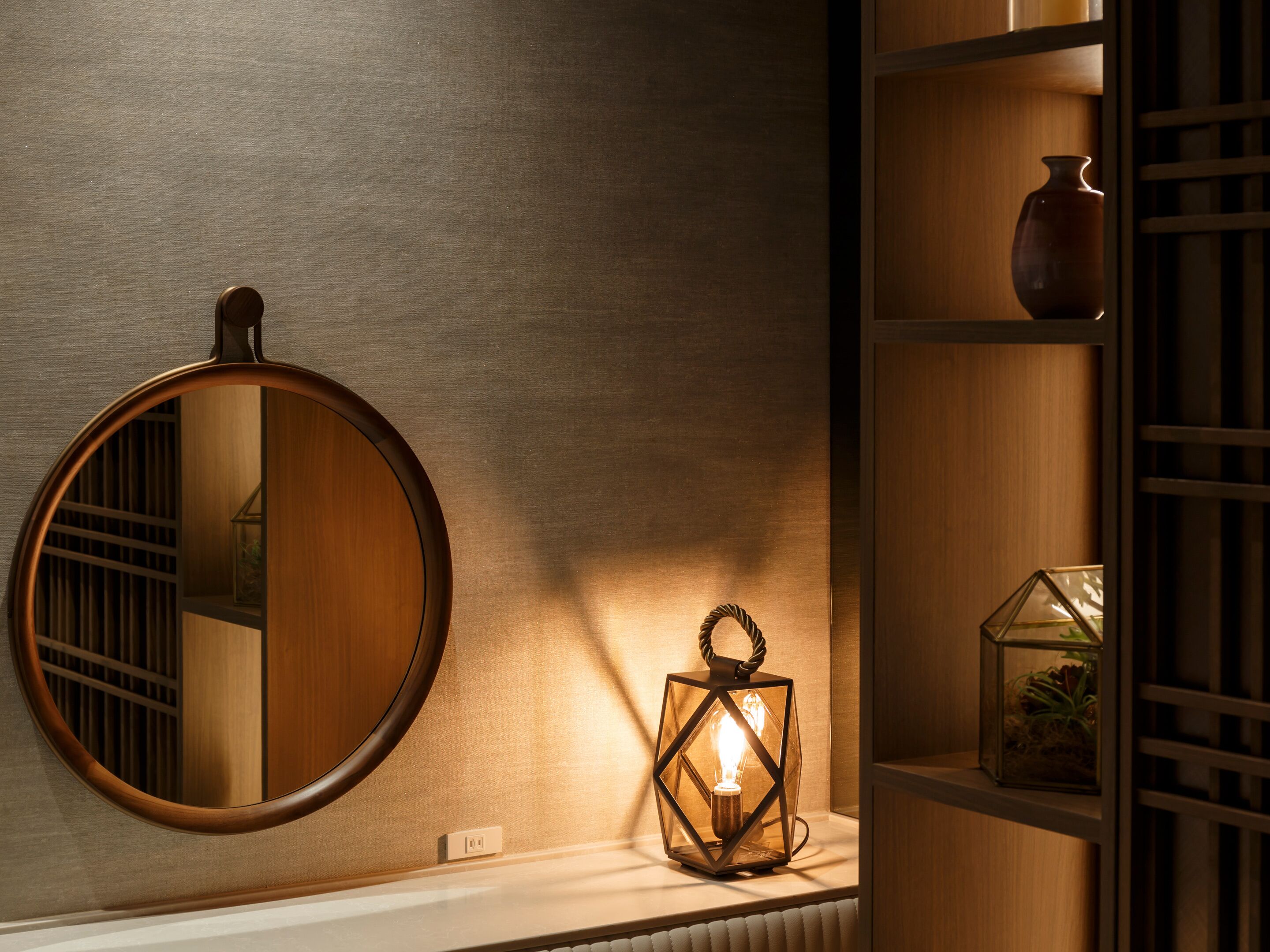 A round mirror, a wall-mounted light fixture, and a shelf with a vase and a glass container.