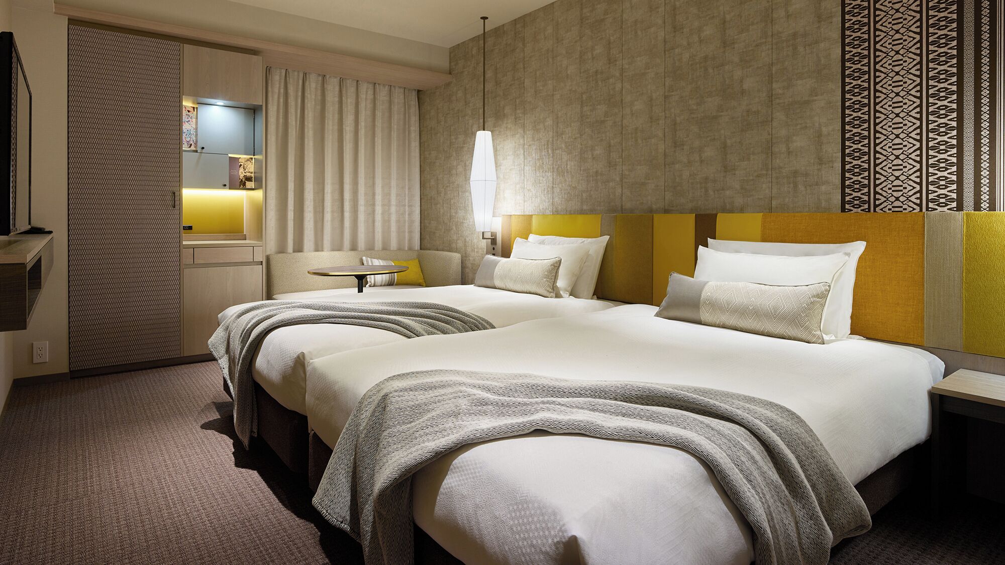 A hotel room with two beds, a bedside table, a lamp, a TV, and a patterned wall.