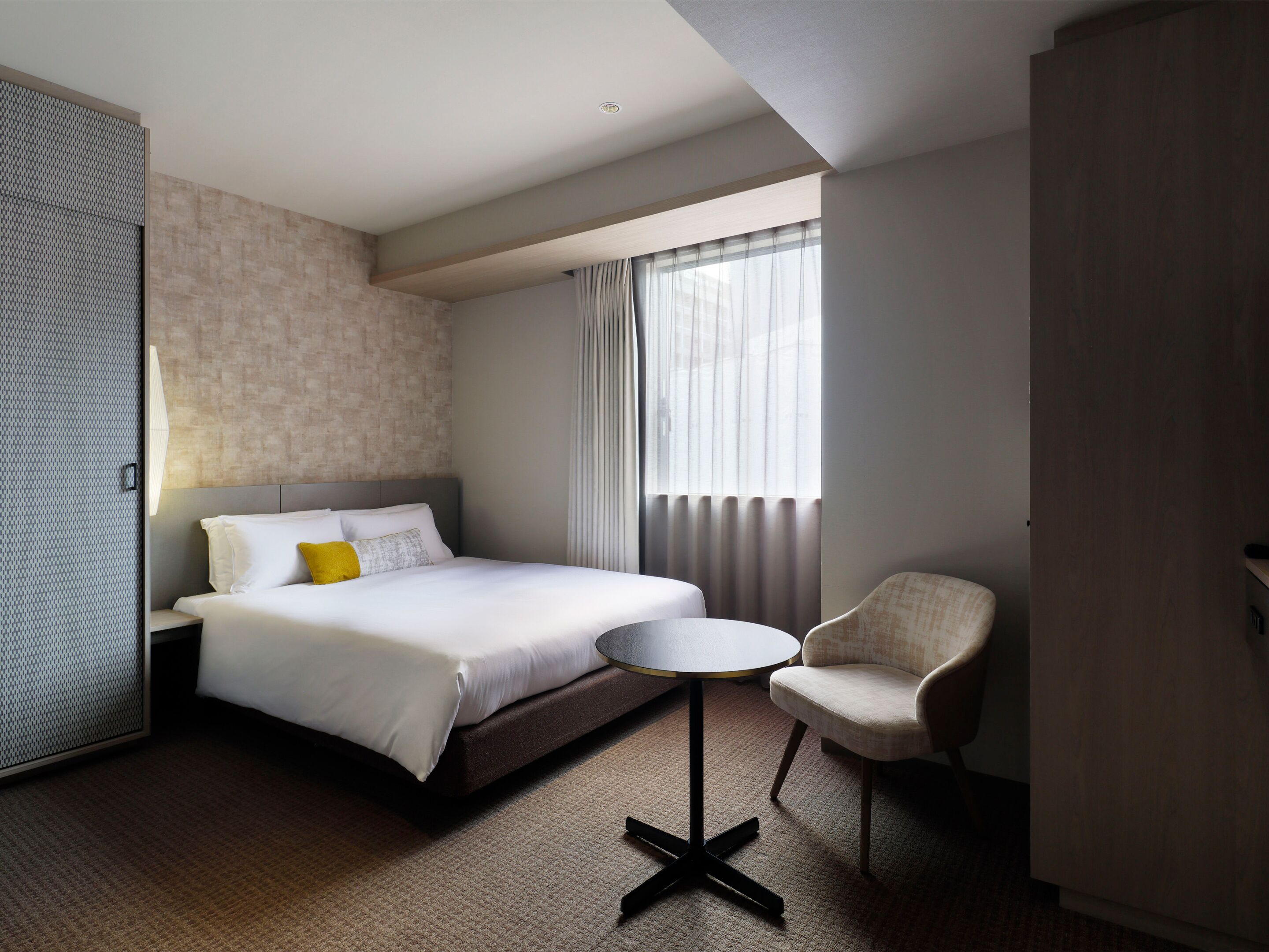 A hotel room with a bed, a small round table, a chair, a wardrobe, and a window with curtains.