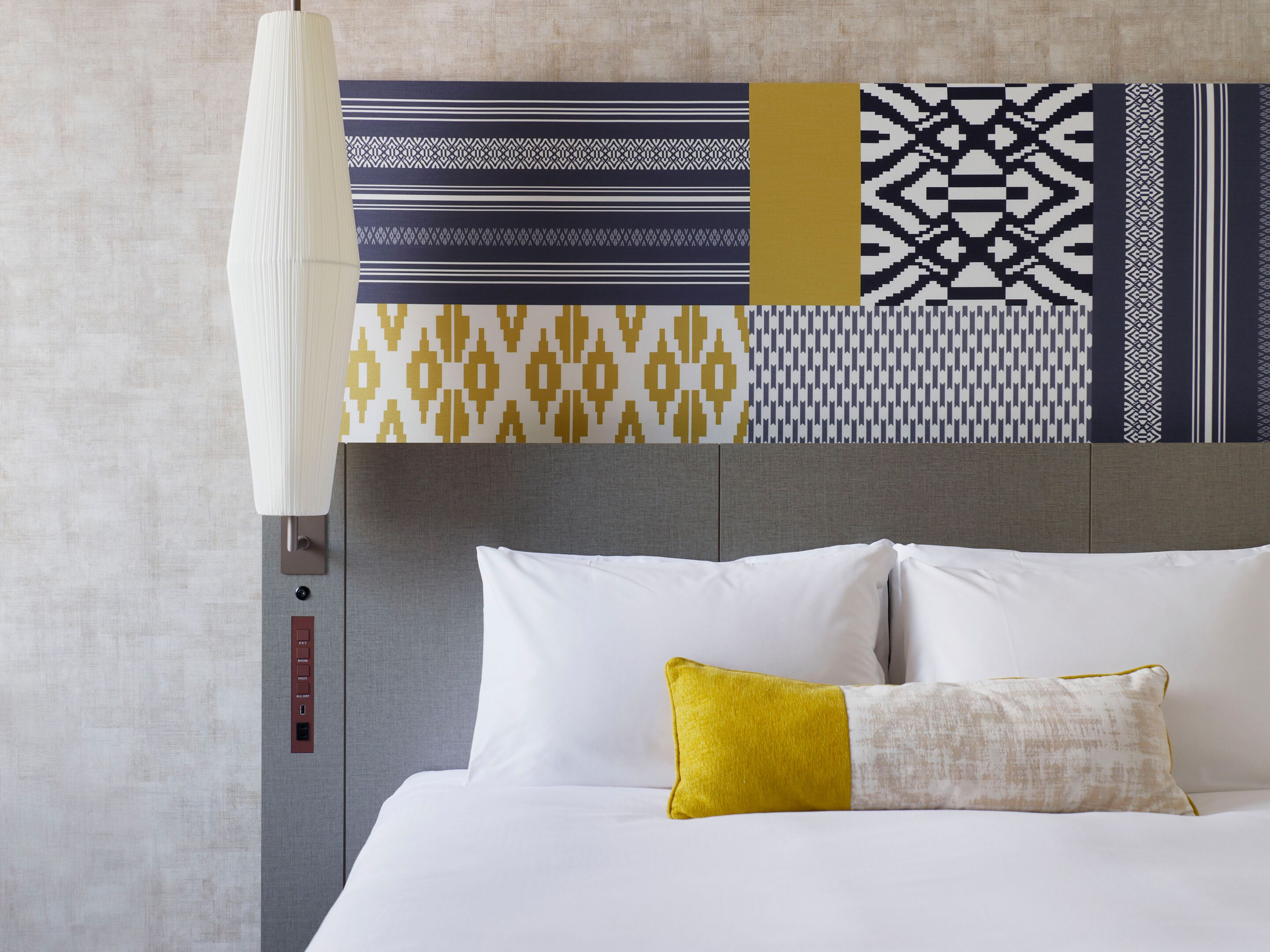 A hotel room with a bed, a headboard, a wall with patterned wallpaper, and a control panel with buttons.