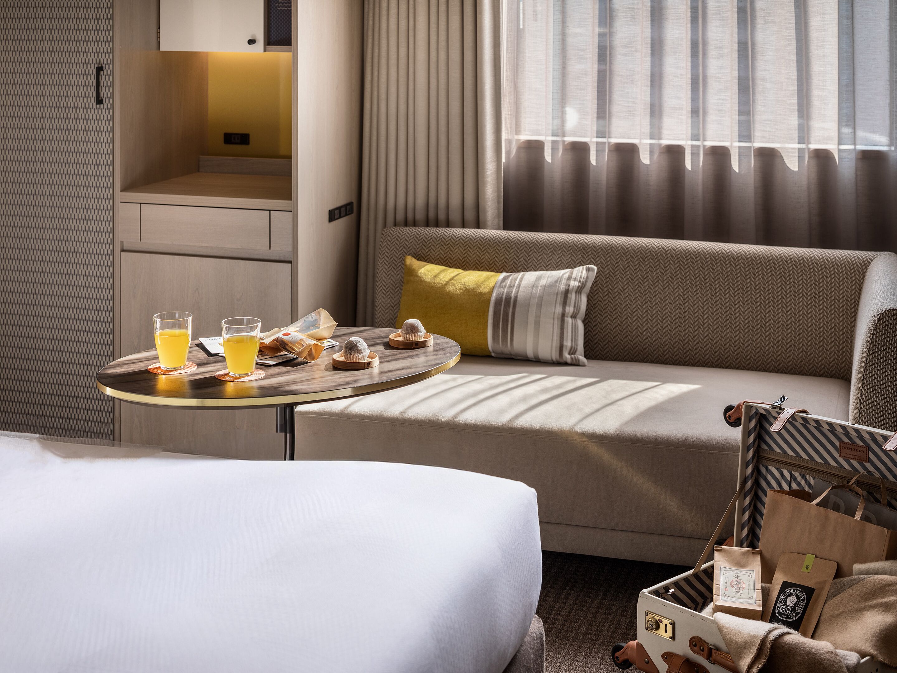 Une chambre d’hôtel avec un canapé, une petite table avec des boissons et des en-cas, un lit et une valise contenant des sacs.