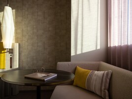 Una habitación moderna con una mesa redonda, un sofá con un cojín estampado y un amplio ventanal con cortinas.