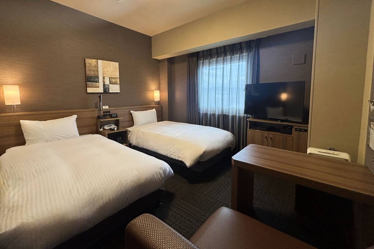 A hotel room with two beds, a desk, a television, and a window with curtains.