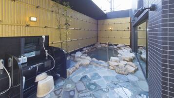 A traditional Japanese onsen with a stone hot spring surrounded by rocks, a black grill, and a bamboo wall.